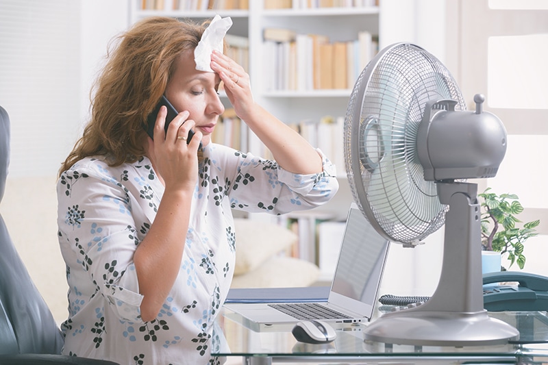 When Do I Replace My Air Conditioner? Woman on the phone attempting to cool herself with a fan.