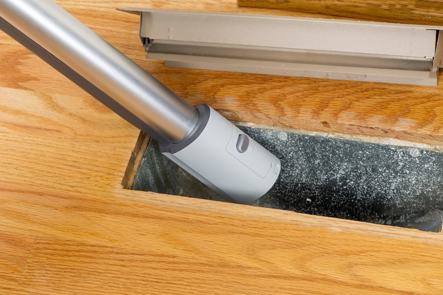 Horizontal photo of vacuum cleaning inside heater floor vent with Red Oak Floors in background. 7 furnace maintenance tips.|Horizontal photo of vacuum cleaning inside heater floor vent with Red Oak Floors in background. 7 furnace maintenance tips.