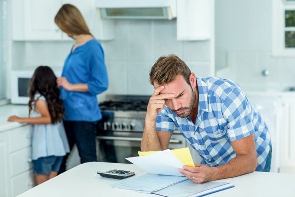 Worried father looking at bills with family in background|Worried father looking at bills with family in background