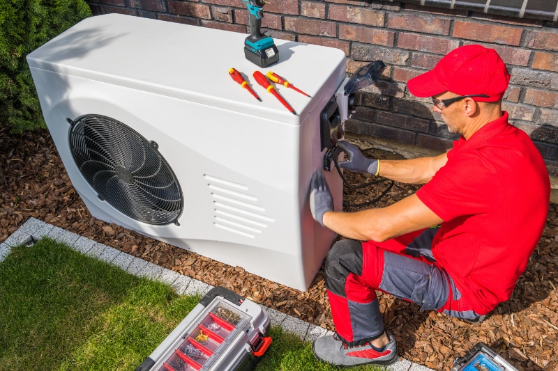 4 Factors to Consider When Buying a Heat Pump. Professional Middle Aged HVAC Technician in Red Uniform Repairing Modern Heat Pump Unit. House Heating and Cooling System Theme.