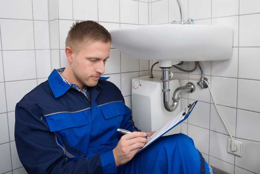 What Questions Should You Ask Before Hiring a Plumber? - Young Plumber Writing On Clipboard With Pen In Kitchen Room.