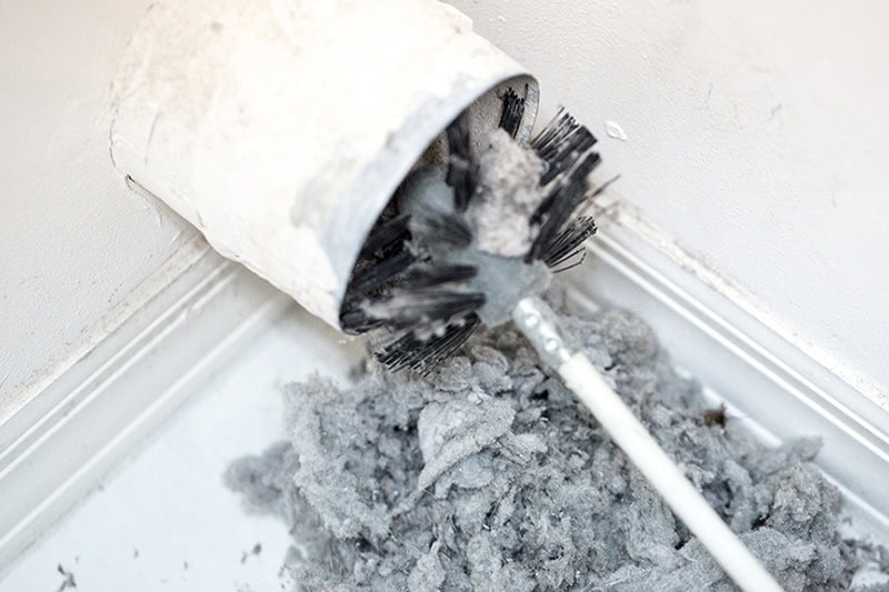 Dryer Vent Cleaning. Dryer vent in a home being cleaned out with a round brush. There is a large pile of lint that has been removed from the vent on a white tiled floor.